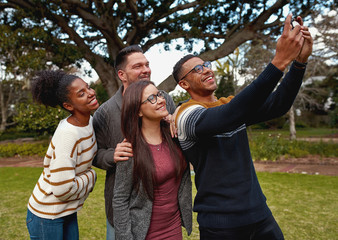 Group of multiracial young friends standing together in a green park taking a selfie on mobile phone smiling - a summer picnic 