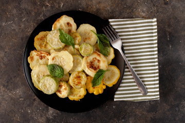 Fried zucchini served with fresh basil leaves. Seasonal vegetable meal, fork and striped napkin on a dark concrete background. Flat lay, close up.