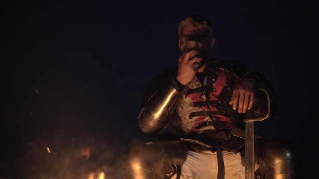 Portrait of medieval knight in armor whit cup sitting near the bonfire.