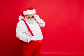 Portrait of handsome old funny fat overweight santa claus with big belly touch his retro specs choose naughty kids wear trousers pants isolated over red background