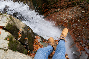 woman in blue jeans and brown boots sitting on the edge looking at waterfall