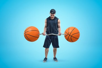 Front view of young athlete in black cap, sleeveless hoodie and shorts, standing and looking down at barbell in his hands, with two basketballs instead of weight plates.