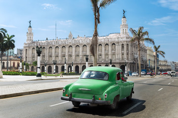 Pphotograph of a vintage green car on the street of downtown Havana: in the background blue sky with few clouds and the tipical palms of Cuba