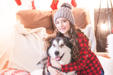 Cute girl in a red checkered shirt and a knitted hat with malamute at home in the room decorated for Christmas. Happy christmas mood