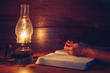 Close up of woman hands hold wooden cross over open bible with oil lamp on wood table while reading bible in the dark room, Christian background devotion or bible study concept