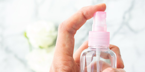 Woman's hand spritzing liquid from small pink spray bottle with marble table and flowers on it in the background. Natural products for cleaning or body care concept