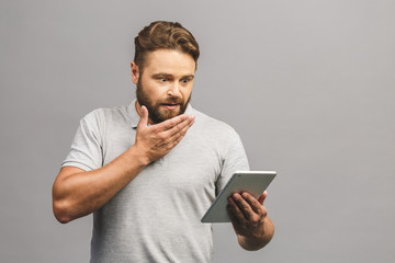 Happy bearded young man in casual standing and using tablet isolated over grey background.
