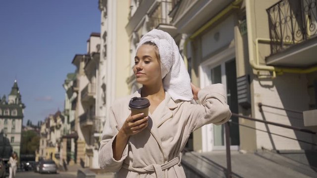 Portrait cute smiling young woman in bathrobe with towel on head walking down the street drinking coffee. Confident girl enjoying a beautiful day in the city
