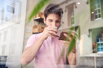 Young Man In Coffee Shop Looking At Mobile Phone Viewed Through Window