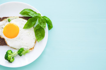 Fried eggs with fresh basil leaves, broccoli and rye bread on bright colorful turquoise backgroud. 
