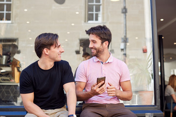 Male Gay Couple Sitting Outside Coffee Shop Looking At Social Media On Mobile Phone