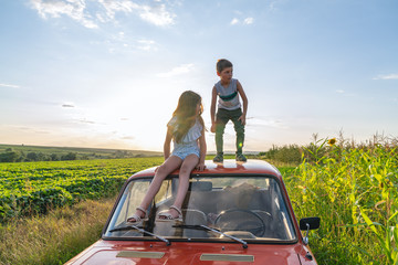Young boy in grey t-shirt standing on the roof of the red retro car and looking scary at the left side her sister in blue striped dress sitting beside and looking at him, field and blue sky on the