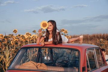 Playful beautiful brunette teen girl in striped blue dress lies on the roof of red retro car and hide one eye with sunflower, blue sky on the background