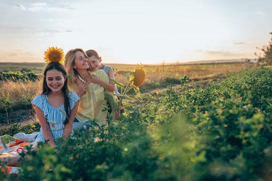 Outdoor photo of beautiful smiling family, teen girl in striped blue dress sitting on plaid among the mealow with sunflower on her head, her brother hugging his happy young mom in yellow shirt