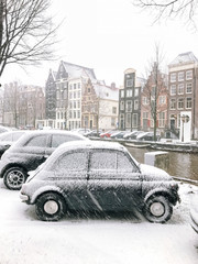 Snowy little car on the bridge in the city center Amsterdam Netherlands. Blizzard on winter in the Netherlands. Small car covered with snow on a bridge. Snowfall in capital Netherlands
