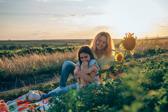 Teen brunette girl sitting calmy at the plaid among the mealow and hugging her legs, her young mom in yellow shirt is sitting beside and looking at ther daughter, sunset on the background