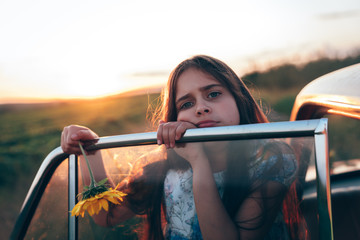 Sad brunette teen girl with sunflower in hand leaned on the car door and looking at the camera, sunset on the background