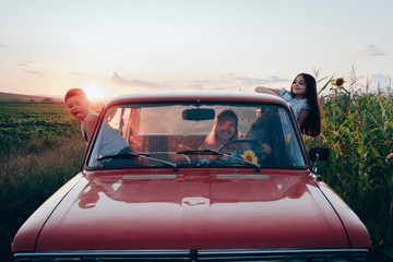 Happy playful family traveling together by the car, beautiful young mom driving and her children got out the car windows and having fun, sunset on the background