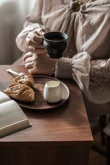 Breakfast on a sunny morning. A cup of coffee in the hands of an adult woman, croissant on the table.