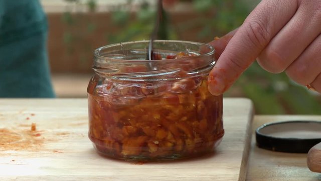 Handheld, close up shot of a person stirring rose hip jam in a glass jar, then placing a lid on top.