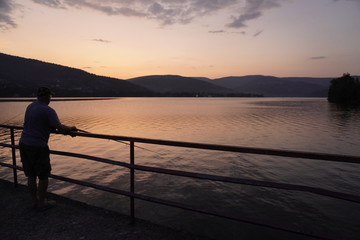 fisherman stands on the pier by the lake, mountainous terrain against the sunset. bright sunset in the mountains. outdoor activity.
