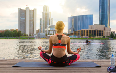 Calm athletic girl meditating at sunset against the background of a pond and passing kayaker. Against evening sky.