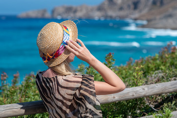 Fototapeta premium Young beautiful European girl in sun hat is standing on mountain beyond wonderful blue sea. She is having her best holidays ever on amazing sea coast.