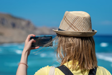 Fototapeta premium Technology, summer holidays, travel and people concept - happy young woman in sun hat taking photo with smartphone over beach background - Image.