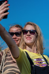 Two young and beautiful female friends in sunglasses are taking selfie on the picturesque sea cost. They are enjoying their summer vacations by sea.