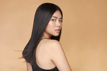 A young beautiful asian girl dressed in a black dress with natural makeup and shiny hair in the wind stands on a beige yellow natural background. Cosmetics concept. Shampoo.
