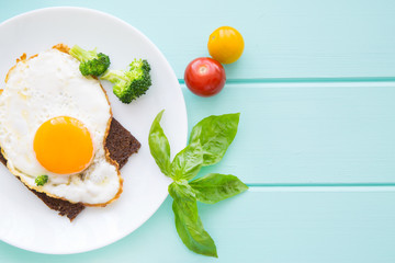 Rye bread toasts with fried eggs and vegetables: cherry tomatoes, broccoli and fresh basil leaves on turquoise table background. Copy space, space for text. 