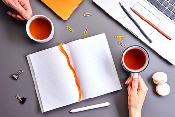 Cups of tea in male and female hands. Laptop, orange book, opened empty notebook and stationery on gray table. Tea break with colleague or friend. Top view with copy space
