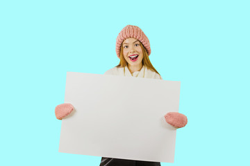 Young woman with blond hair, wearing casual outfit, boots, is sitting on white floor, holding white makeup poster, isolated on white background