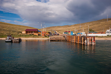 Fototapeta na wymiar Ferry pier Olkhon island with waiting cars and people. On the shore are wooden houses, lampposts. Over the hills clouds.