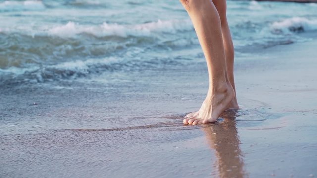 Legs of a young slender ballet dancer girl standing on sandy sea shore. Feet washed by warm waves with foam