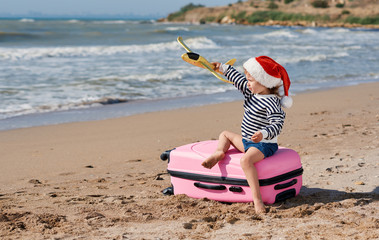 Obraz premium Little girl in Santa hat playing with toy airplane on sunny tropical beach. Child sits on pink suitcase. Christmas and new year vacation travel concept