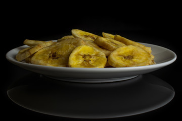 Lot of slices of sweet yellow dry banana on white ceramic plate isolated on black glass
