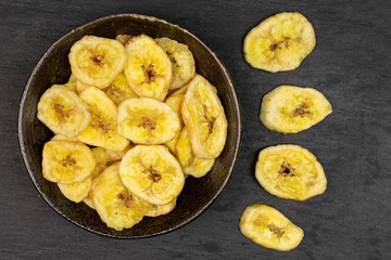Lot of slices of sweet yellow dry banana in dark ceramic bowl flatlay on grey stone