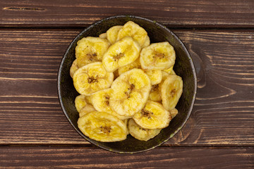 Lot of slices of sweet yellow dry banana in dark ceramic bowl flatlay on brown wood
