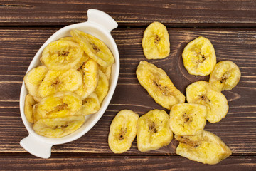 Lot of slices of sweet yellow dry banana in white oval ceramic bowl flatlay on brown wood