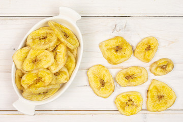 Lot of slices of sweet yellow dry banana in white oval ceramic bowl flatlay on white wood