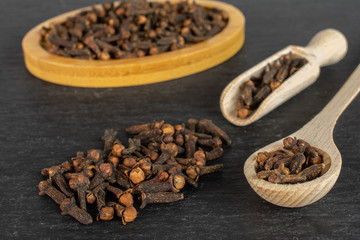 Lot of whole dry brown clove on bamboo coaster in a wooden scoop in a wooden spoon on grey stone