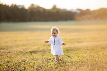 Little curly, blue-eyed girl in a white dress walks in the field. Portrait of a little girl in the sun. Sunset.