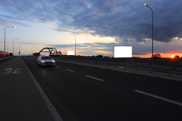 Samochód osbowy na drodze, zachód słońca, bilbord reklamowy, tekst, wiadukt, most.	