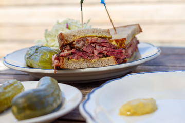 Pastrami sandwich sliced in half on rye bread with mustard on a white plate on a wood table as seen from above. Coleslaw and dill pickle on side. Toothpicks in both halves of sandwich. 