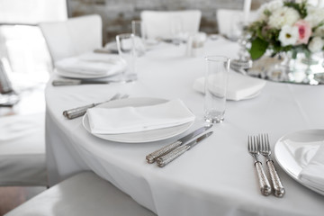 Wedding table setting decorated with fresh flowers. White plates, silverware, white tablecloth and white room. Wedding floristry. Banquet table for guests. Bouquet with roses, hydrangea and eustoma