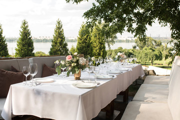 Wedding table setting decorated with fresh flowers in a brass vase. Wedding floristry. Banquet table for guests outdoors with a view of green nature. Bouquet with roses, eustoma and eucalyptus leaves