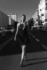 Vertical black and white photo of full body view of elegant woman, in classic black dress and high heels. Walking by the street with city buildings and cars in the background.