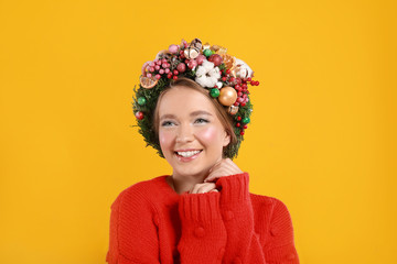 Beautiful young woman wearing Christmas wreath on yellow background