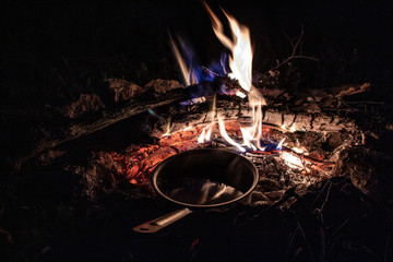 A pot of water is heated by the fire. Tourist bonfire at night in the forest. The flame of fire warms a ladle with water. Boiling water in the wild.